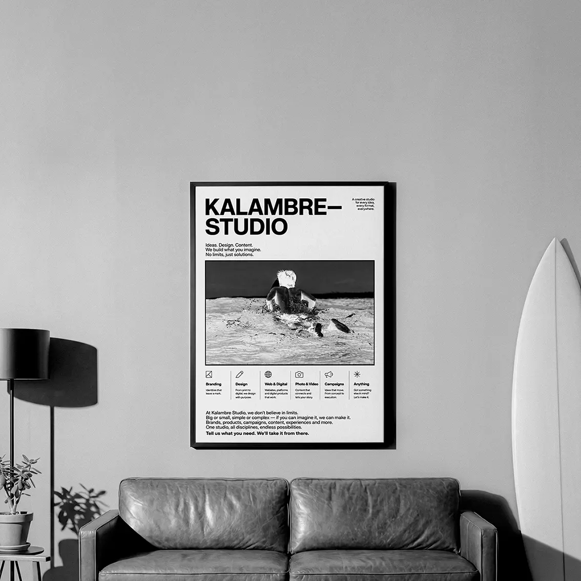 Framed Kalambre Studio poster with a black and white surf photo hung in a living room with a leather sofa and a surfboard beside it
