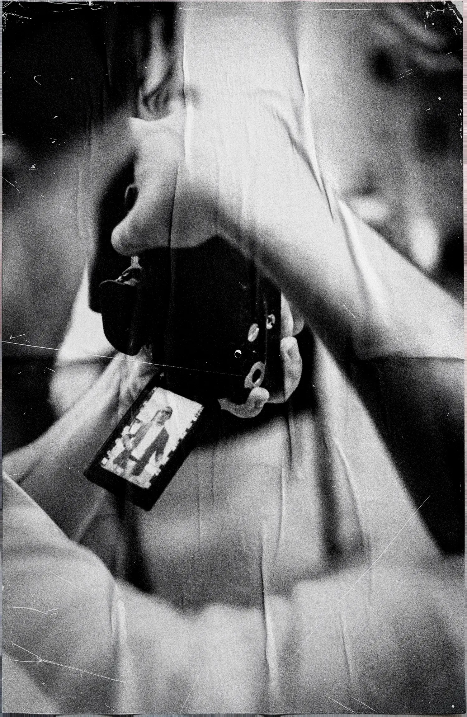 Black and white backstage detail of a shoot: hands hold a camera with a developed film frame hanging from it, over a crumpled-paper texture.