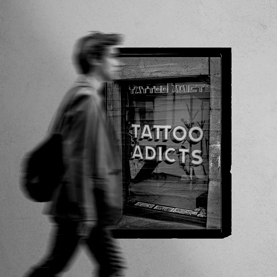 Black and white street photograph of a man with a bag walking, motion-blurred, past the Tattoo Adicts shop window